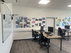 Blick in ein Büro mit drei Schreibtischen auf denen jeweils Bildschirme, Tastatur und Maus zu sehen sind. An den Wänden viele Bilder sowie ein Whiteboard.