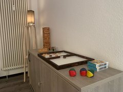 Auf einem Sideboard, das an der Wand neben einer eingeschalteten Stehlampe steht, stehen diverse Spiele, wie z. B. Jenga, Jonglierbälle etc. zur Verfügung.