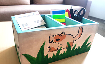 Schreibtisch-Utensilo aus Holz mit drei Fächern, in denen Blöcke, Stifte, eine Schere sowie PostIts zu sehen sind. Bemalt ist die Kiste mit einer grünen Wiese auf der eine braun-weiße Katze sitzt. 