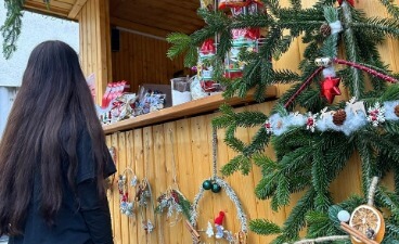 Zu sehen ist eine dunkel gekleidete Frau mit langen dunklen Haaren, die vor eine mit Tannenzweigen und Weihnachtsschmuck geschmückten Weihnachtshütte steht.